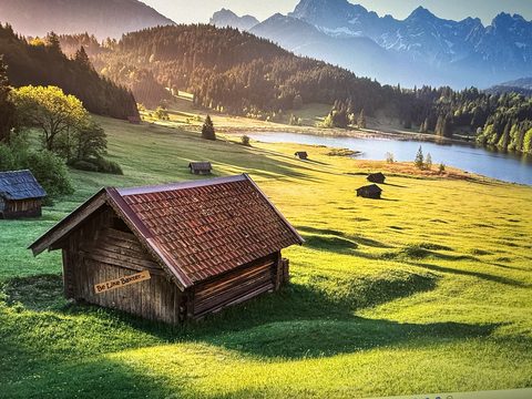 A Be Like Baxter bumper sticker on the wall of an old wooden barn in a green alpine valley, with mountains and a lake behind it