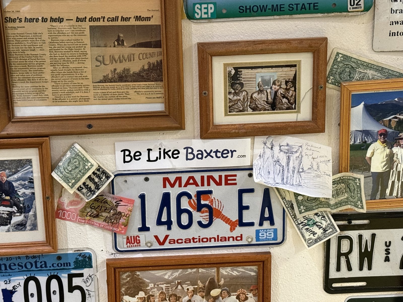 A wall crowded with licence plates, framed photographs and clippings from many different places, with a Be Like Baxter sticker among them