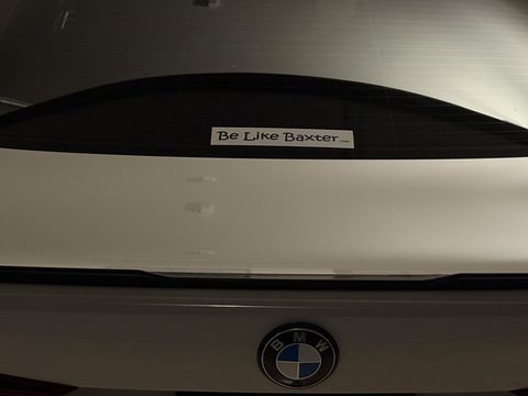 A Be Like Baxter bumper sticker on the rear window of a white car in a garage