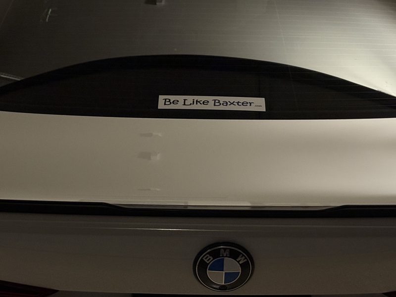 A Be Like Baxter bumper sticker on the rear window of a white car in a garage