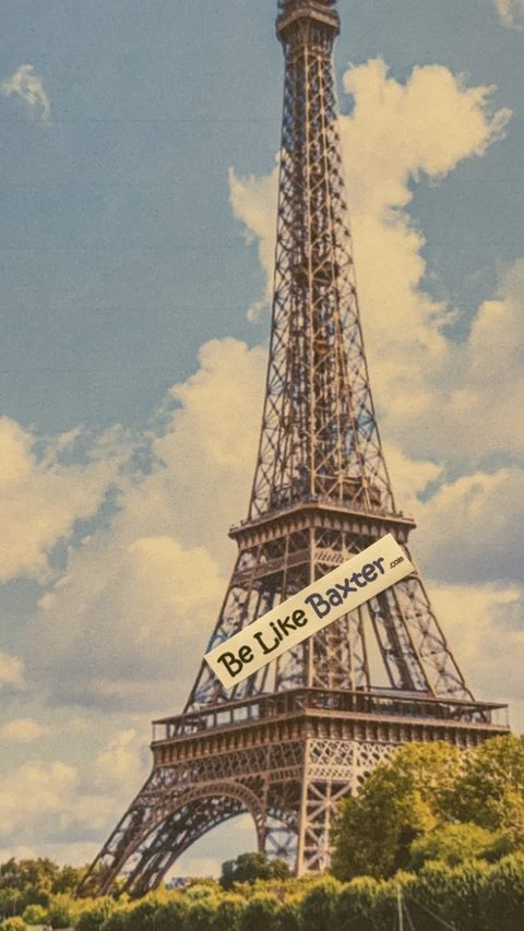 A Be Like Baxter bumper sticker laid diagonally across a vintage print of the Eiffel Tower
