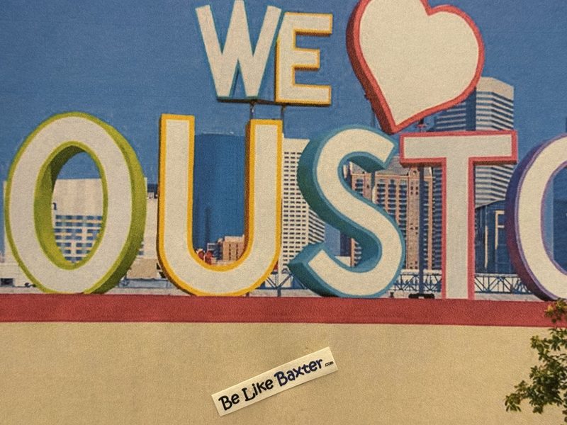 A Be Like Baxter bumper sticker resting against a large outdoor mural of colourful letters spelling WE LOVE HOUSTON