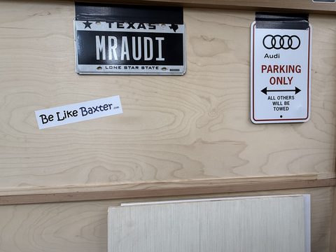 A Be Like Baxter bumper sticker taped to a plywood wall beneath a Texas vanity licence plate reading MRAUDI and an Audi Parking Only sign