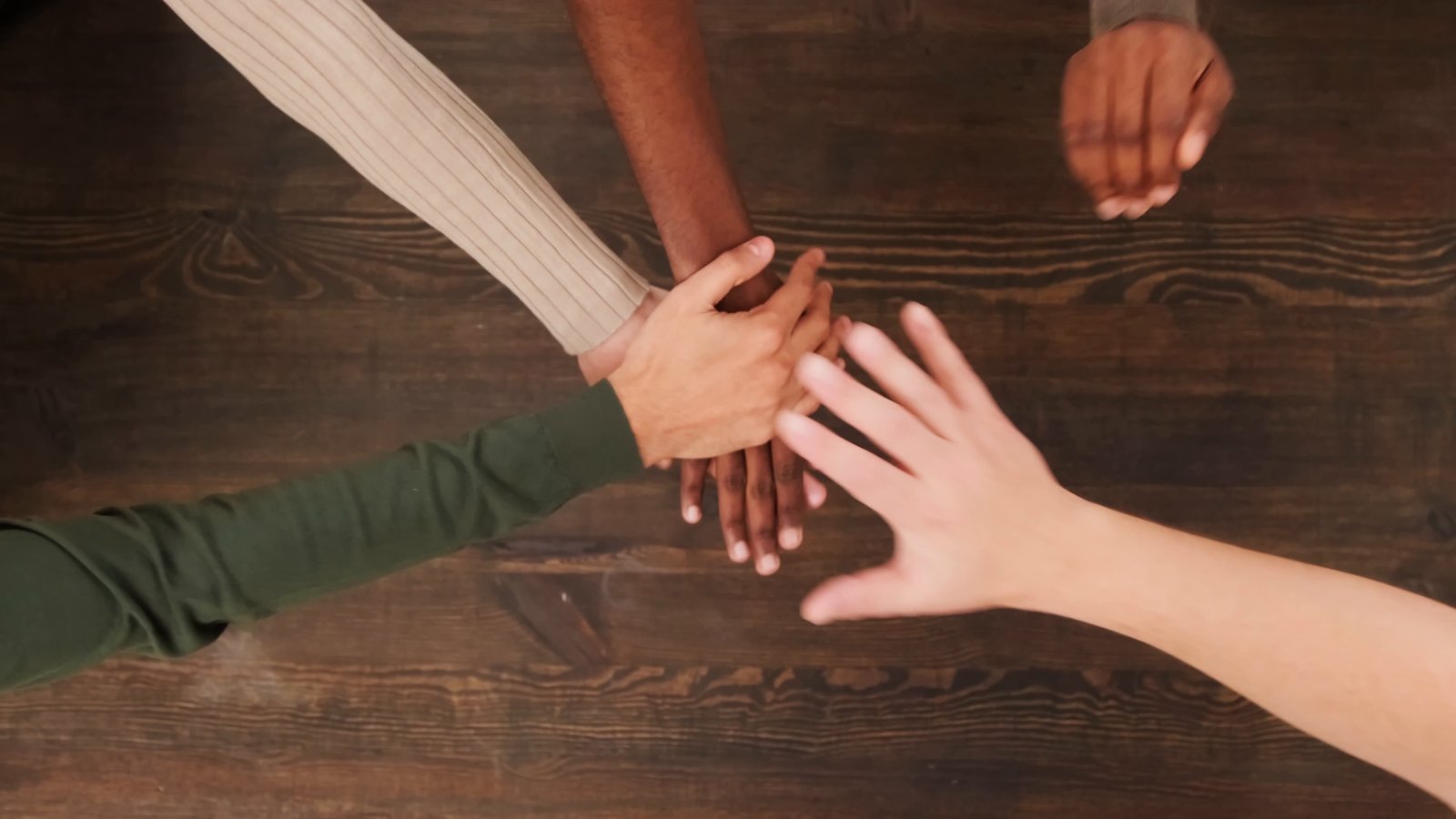 An overhead shot of hands with different skin tones stacking together on a wooden table